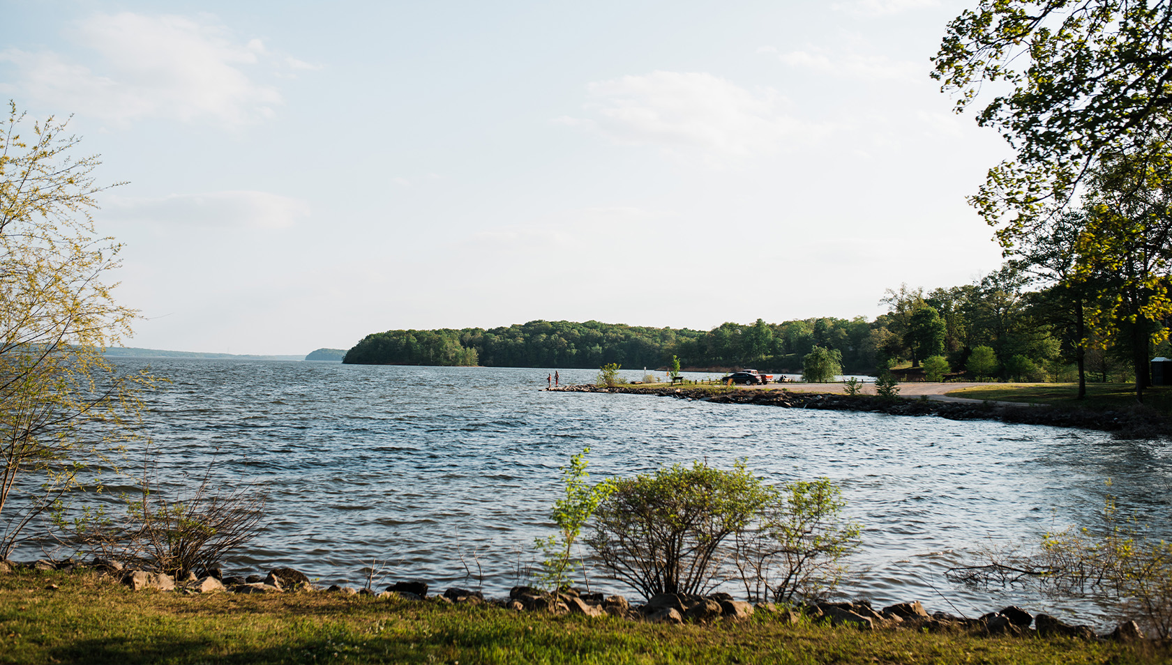 A scenic lakeside view at Paris Landing State Park with gentle waves, a distant tree-covered shoreline, and a small jetty where a few people stand near parked cars under a clear sky.