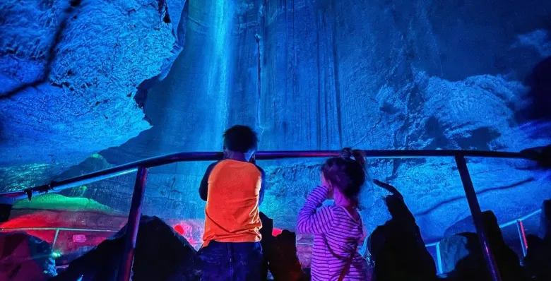 Visitors viewing a tall underground waterfall illuminated by blue and purple lights.